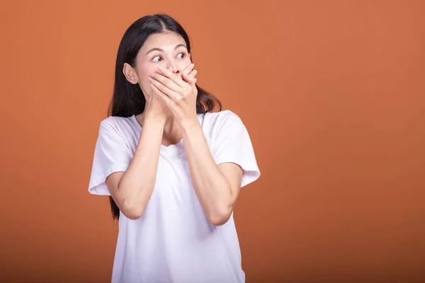 Surprise woman over orange background. Young Asian hipster woman in white t-shirt with very surprise pose, two hands cover mouth. Young woman hipster concept.