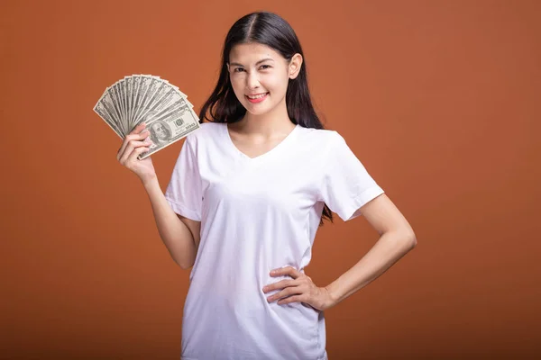 Woman holding cash notes isolated in orange background. Young asian woman in white t-shirt in winning pose, holding dollar note. Young rich hipster concept.