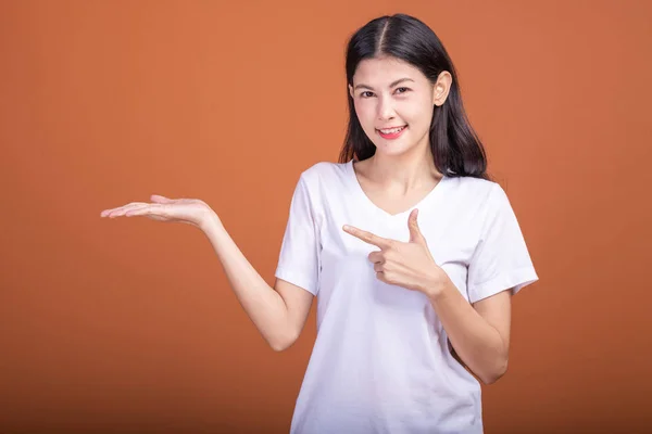 Woman pointing finger with open palm over orange background. Young Asian hipster woman in white t-shirt posing open palm with happy emotion.