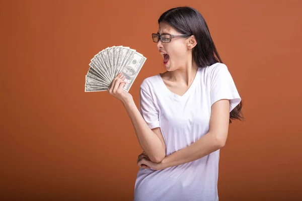 Woman holding cash notes isolated in orange background. Young asian woman in white t-shirt, nerd glasses in very confident win mood, holding dollar note. Young rich hipster concept.