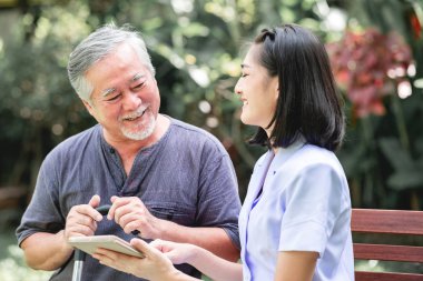 Hemşire hasta birlikte tablet vasıl seyir bankta oturmuş ile. Asyalı yaşlı adam ve birlikte konuşurken oturan bir genç bayan. Ruh sakin ol.