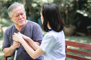 Hemşire ile hasta birlikte konuşurken bankta oturan. Asyalı yaşlı adam ve birlikte konuşurken oturan bir genç bayan. Ciddi ruh hali.