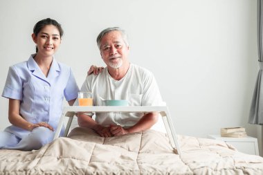Young attractive nurse and senior man in bed portrait. Old asian male with white beard with young attractive asian woman caretaker. Senior home service concept. clipart