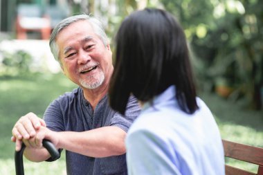 Hemşire ile hasta birlikte konuşurken bankta oturan. Asyalı yaşlı adam ve birlikte konuşurken oturan bir genç bayan. Mutlu bir gülümseme.