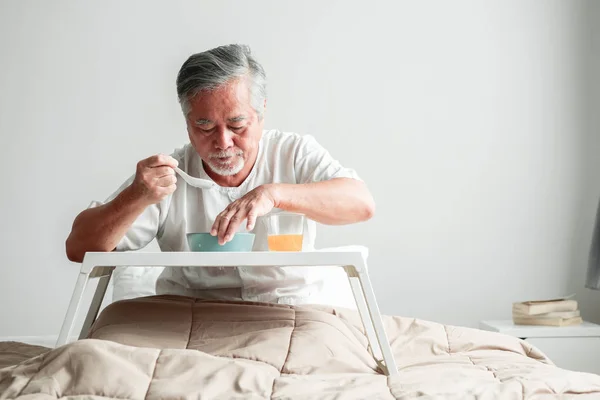 Senior man in bed enjoying breakfast. Old asian male with white beard eating congee and orange juice. Senior home service concept.