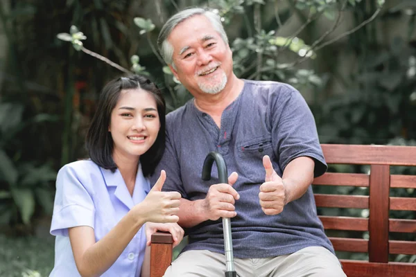 Nurse with patient sitting on bench together with thumbs up. Asian old man and young woman sitting together talking. Very happy mood.