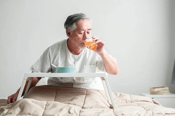 Senior man in bed enjoying breakfast. Old asian male with white beard drinking orange juice. Senior home service concept.