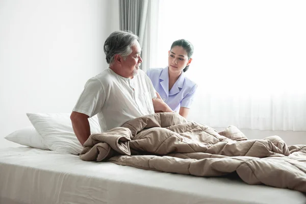 Senior man in bed trying to get up and nurse helping him. Old asian man and beautiful asian nurse woman in bedroom and open curtain. Senior home service concept.