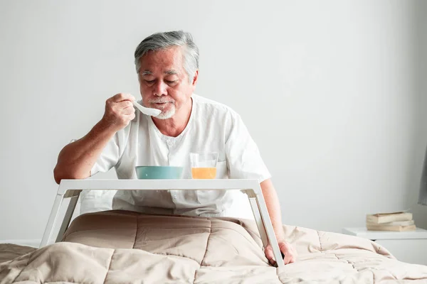 Senior man in bed enjoying breakfast. Old asian male with white beard eating congee and orange juice. Senior home service concept.