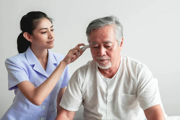 Beautiful nurse using high tech Temperature measurement tool on old man. Senior Asian man with white beard in bed with attractive asian woman nurse. Senior retirement home service, care taker concept.