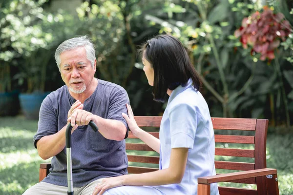 Nurse with patient sitting on bench together talking. Asian old man and young woman sitting together talking. Serious mood.