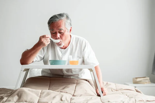 Senior man in bed enjoying breakfast. Old asian male with white beard eating congee and orange juice. Senior home service concept.