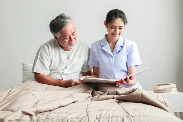 Young beautiful female nurse reading book to senior man. Asian woman reading book to old asian man with white beard in bed.