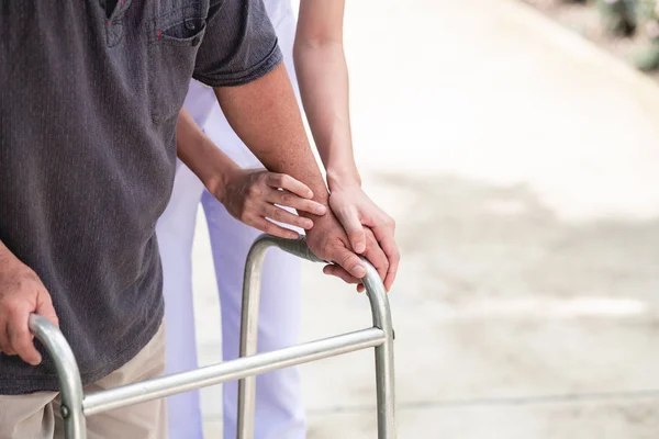 Nurse with patient using walker in retirement home. Young female nurse holding old man\'s hand in outdoor garden walking. Senior care, care taker and senior retirement home service concept.