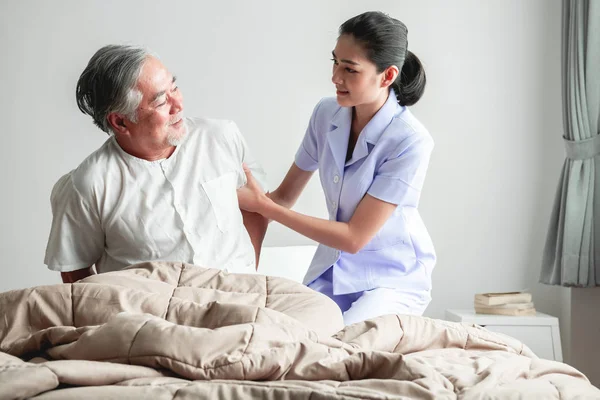 老人在床上试图起床和护士帮助他 老亚裔男子和美丽的亚洲护士妇女在卧室和开放窗帘 高级家庭服务理念 — 图库照片