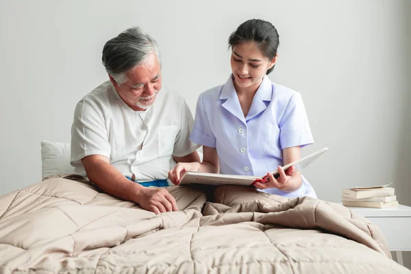 Young beautiful female nurse reading book to senior man. Asian woman reading book to old asian man with white beard in bed.