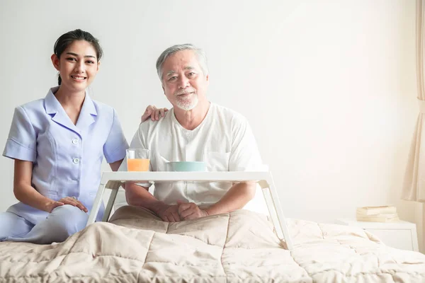 Young attractive nurse and senior man in bed portrait. Old asian male with white beard with young attractive asian woman caretaker. Senior home service concept.