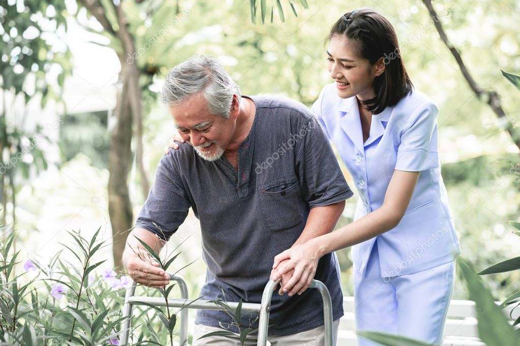 Nurse with patient using walker in retirement home. Young female nurse holding old man's hand in outdoor garden walking. Senior care, care taker and senior retirement home service concept.