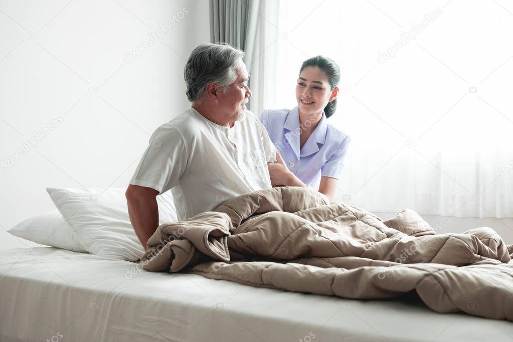 Senior man in bed trying to get up and nurse helping him. Old asian man and beautiful asian nurse woman in bedroom and open curtain. Senior home service concept.
