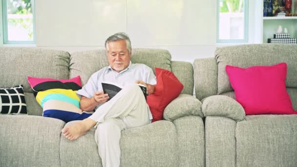 成熟的亚洲人穿着白色坐在沙发上看书和微笑的室内 — 图库视频影像
