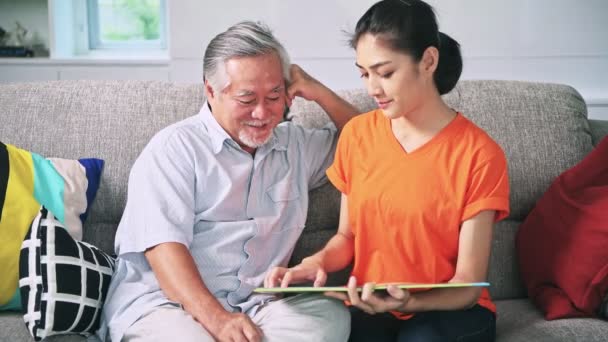 亚洲祖父与灰色的头发和孙女坐在沙发上微笑的谈话和阅读书 — 图库视频影像