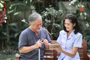 Hemşire hasta birlikte tablet vasıl seyir bankta oturmuş ile. Asyalı yaşlı adam ve birlikte konuşurken oturan bir genç bayan. Ruh sakin ol.