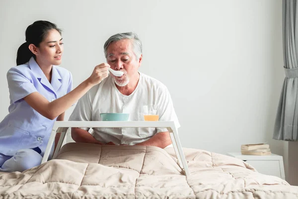 Young attractive nurse feeding breakfast to senior man in bed. Old asian male with white beard with young attractive asian woman caretaker. Senior home service concept.