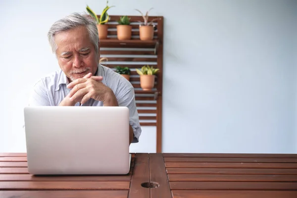 Old man trying to use computer in outdoor balcony. Asian man with white beard using laptop, upset mood. Senior lifestyle concept.