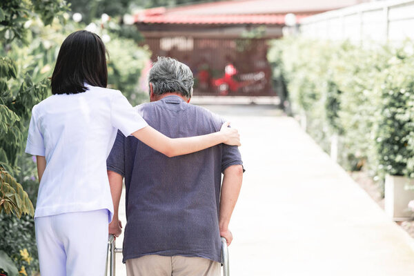Nurse with patient using walker in retirement home. Young female nurse holding old man's shoulder in outdoor garden walking. Senior care, care taker and senior retirement home service concept.