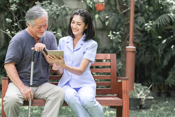 Nurse with patient sitting on bench together looking at tablet. Asian old man and young woman sitting together talking. Relax, happy smile mood.