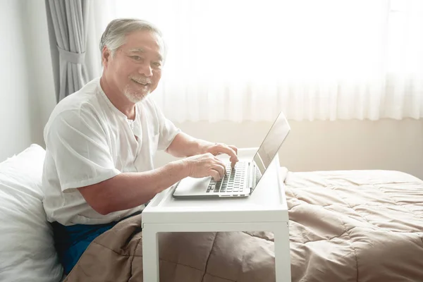 Old man typing on key board in bed. Senior asian man in bed working on laptop.