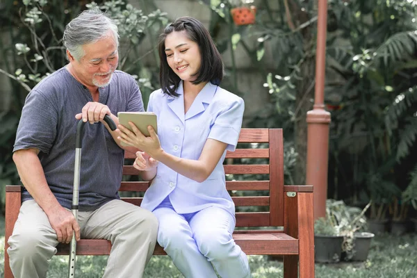 Nurse with patient sitting on bench together looking at tablet. Asian old man and young woman sitting together talking. Relax, happy smile mood.