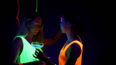 Güzel seksi kadın UV boya, lazer, parlak bilezik, İçecek, giyim birlikte yarım vücut shot kamera önünde dans parlayan yüz. Beyaz ve Asyalı bir kadın. Parti konsept.