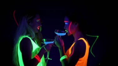 Güzel seksi kadın UV boya, lazer, parlak bilezik, İçecek, giyim birlikte yarım vücut shot kamera önünde dans parlayan yüz. Beyaz ve Asyalı bir kadın. Parti konsept.