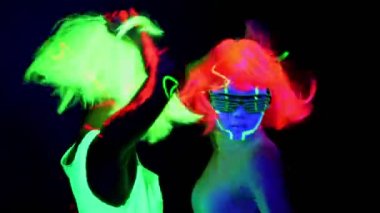 Gözlük, giyim, birlikte hızlı kamera önünde, yarım vücut vurdu dans parlayan parlayan güzel seksi kadın Uv yüz boyama, peruk, ile. Beyaz ve Asyalı bir kadın. Parti konsept.