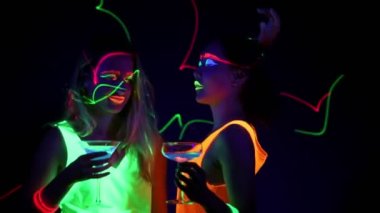 Güzel seksi kadın UV boya, lazer, parlak bilezik, İçecek, giyim birlikte yarım vücut shot kamera önünde dans parlayan yüz. Beyaz ve Asyalı bir kadın. Parti konsept. 