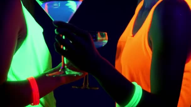 紫外線で美しいセクシーな女性顔ペイント 光るブレスレット 飲み物 衣類を閉じるカメラの前でドリンクの一緒に踊って光る 白人とアジアの女性 パーティー コンセプト — ストック動画