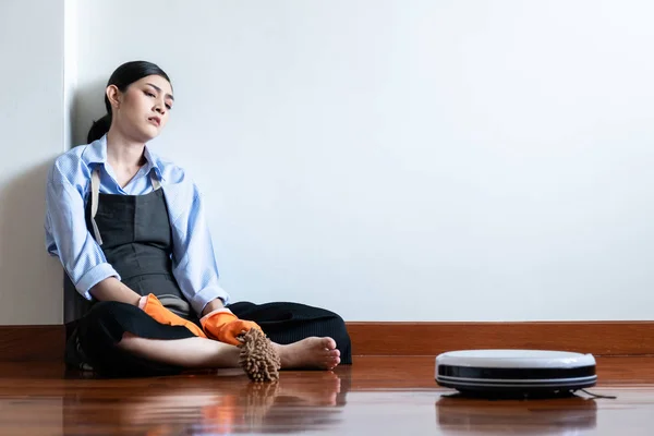 Tired house wife sitting on floor with cleaning robot passing by. Beautiful Asian woman sitting on floor looking at cleaning robot. Robot autonomus cleaning service concept.