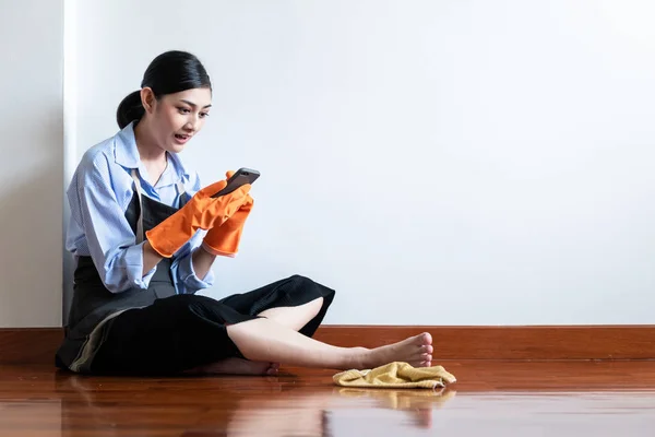 疲惫的家庭主妇在地板上休息 美丽的亚洲妇女坐在地板上休息与旧布和发现清洁服务在她的手机 低角度拍摄 网上房屋清洁服务概念 图库照片