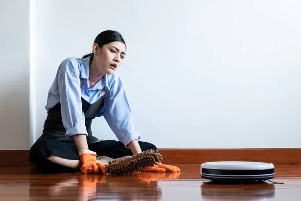 Tired house wife sitting on floor with cleaning robot passing by. Beautiful Asian woman sitting on floor looking at cleaning robot. Robot autonomus cleaning service concept.