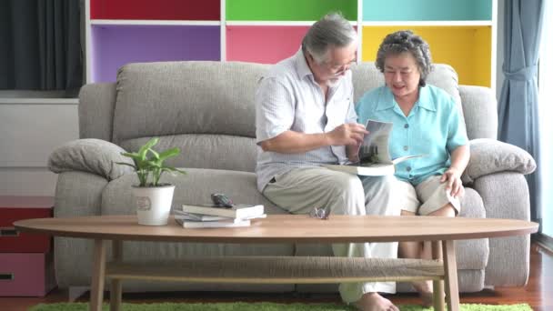 シニア カップルに座って 読書をリビング ルームで一緒に予約します 引退した古いアジア男性と女性 一緒に幸せな笑顔を本を読みます シニアのライフ スタイルのコンセプト — ストック動画