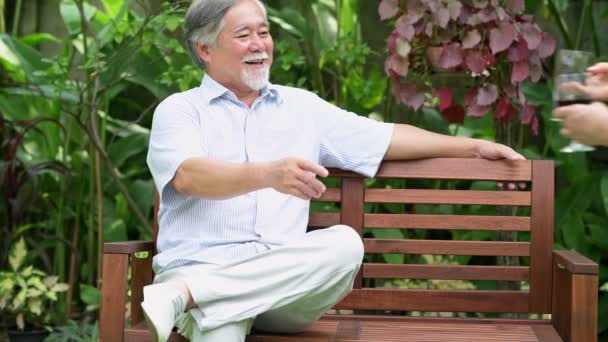 年配のカップルが座っていると 家の庭で一緒に赤ワインを飲みます 引退した古いアジアの男性と女性 幸せな笑顔にワイングラスをもたらします シニアのライフ スタイルのコンセプト — ストック動画