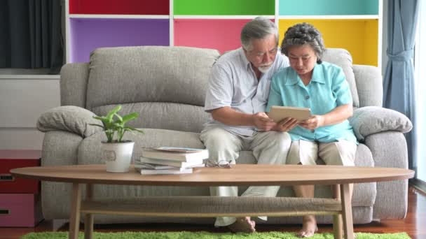 资深夫妇坐在客厅里看平板电脑 退休的亚洲男女老人 喜欢在客厅沙发上玩平板电脑 开心的微笑 老年人生活方式理念 — 图库视频影像