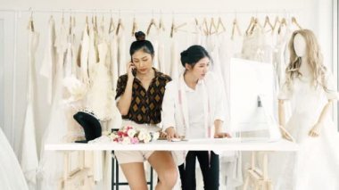 Birlikte çalışan genç moda evi tasarımcı. Bir gelinliğin genç Asyalı kadın ile müşteriler emir alan bilgisayar hem de telefonda. Küçük işletme kavramı.