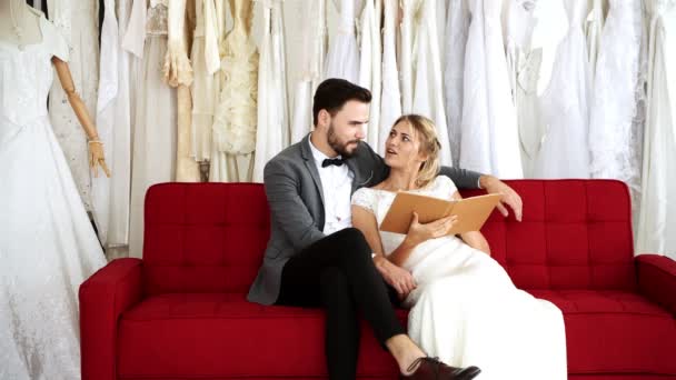 高価なドレス ショップでロマンチックな結婚式です 白人男性と女性の花嫁ドレス 本を見ているソファーに一緒に座っていた 若いカップルのロマンチックな概念 — ストック動画