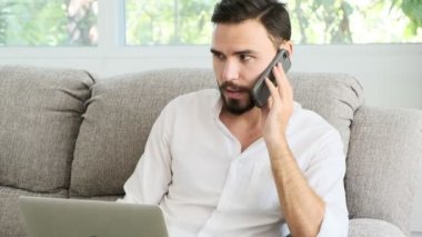 Onun dizüstü bilgisayarda çalışan ve müşteri için telefonda konuşurken kanepede oturan genç adam. Sakal, freelence kavramı ile beyaz erkek.
