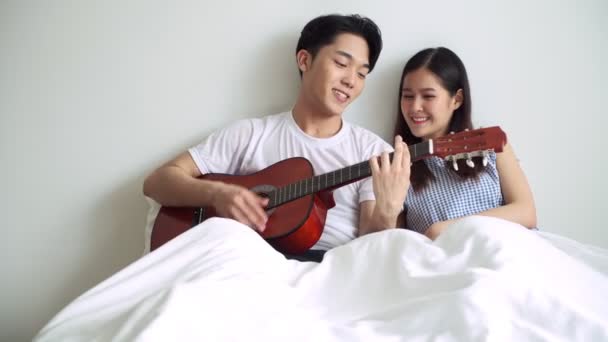 彼のガール フレンドを喜んでギターを弾く男とベッドでカップル アジア中国男と一日の時間でベッドの女 現代の夫婦のライフ スタイル コンセプト — ストック動画