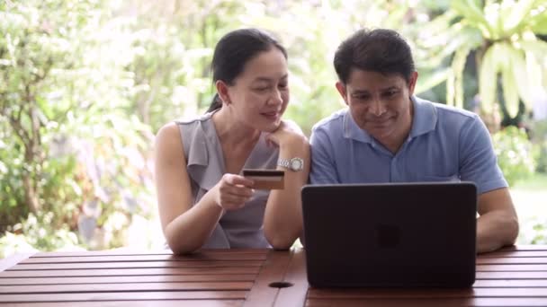 亚洲高级男子试图在信用卡号码 他的妻子拿着到笔记本电脑户外与美丽的热带花园的背景 高级网上购物理念 — 图库视频影像