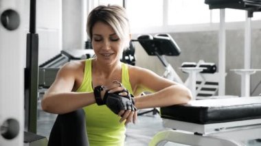 Beyaz tenli sarışın kadın spor salonunda akıllı bir saat kullanıyor. Bankta oturuyor, saatine bakıyor..