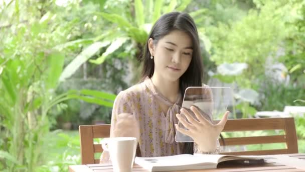 若い美しいアジアの女性が背景に緑の木と彼女の家の庭でハイテクガラスディスプレイで彼女のソーシャルメディアフィードを閲覧し 幸せな笑顔 将来の技術コンセプト グラフィックオーバーレイの場合 — ストック動画